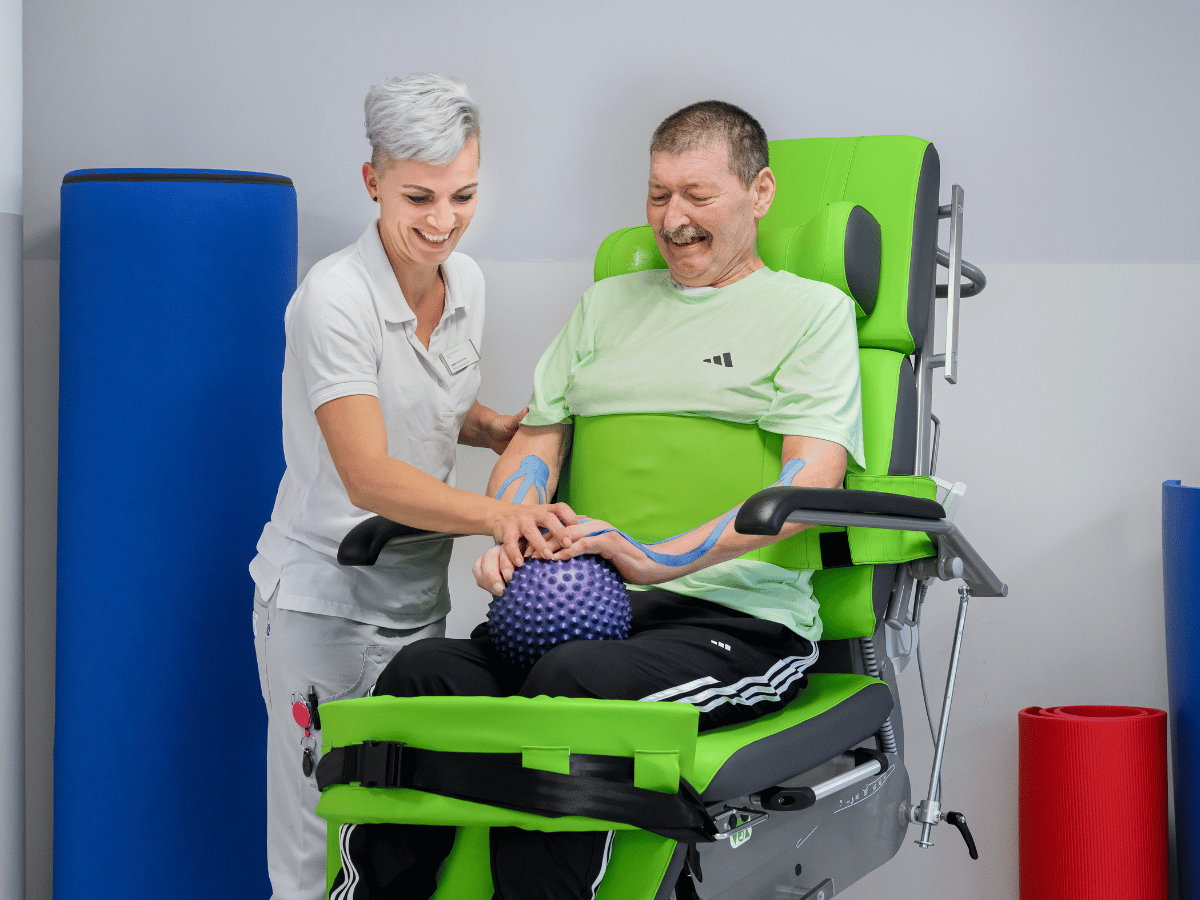 Die Patientin sitzt auf dem Stuhl mit einem lila Ball und neben ihr die Pflegerin 