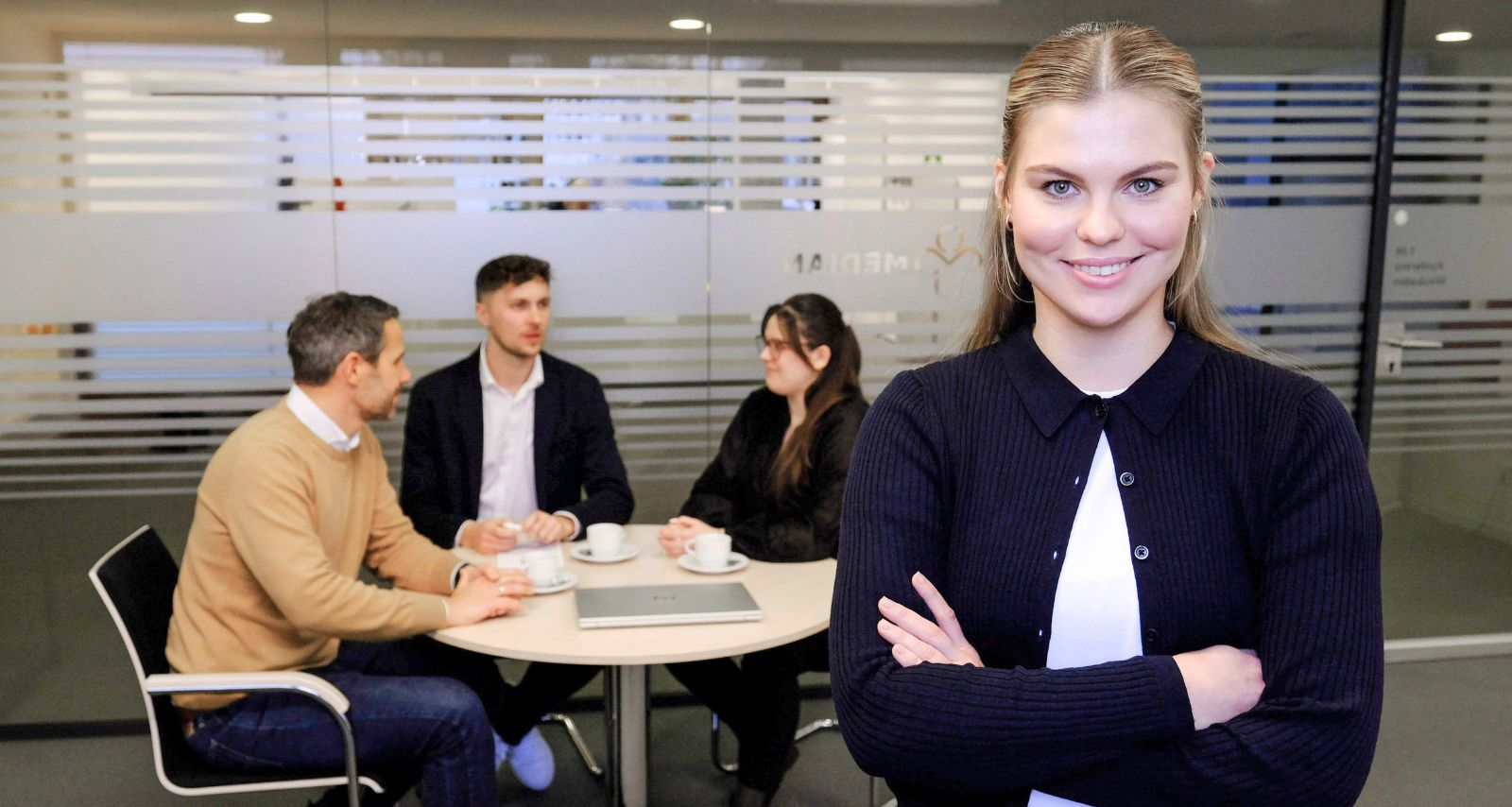 Eine Frau die lächelt und hinter ihr sitzen drei Personen die sich unterhalten