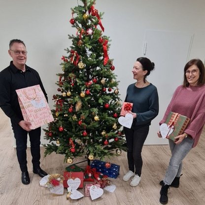 Drei Personen die Geschenke halten, am Baum stehen und die Kamera lächeln 