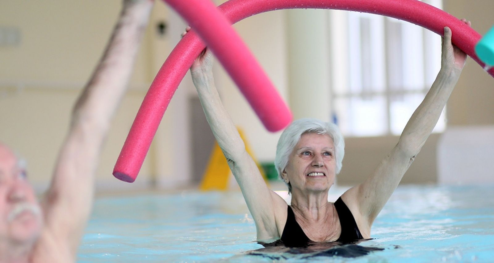 Patientin streckt eine pinke Poolnudel über den Kopf und lächelt bei der Wassergymnastik