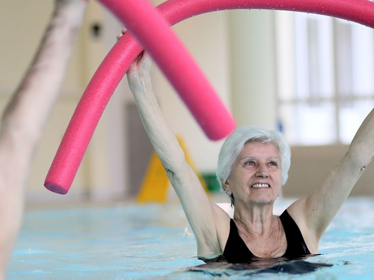 Patientin streckt eine pinke Poolnudel über den Kopf und lächelt bei der Wassergymnastik