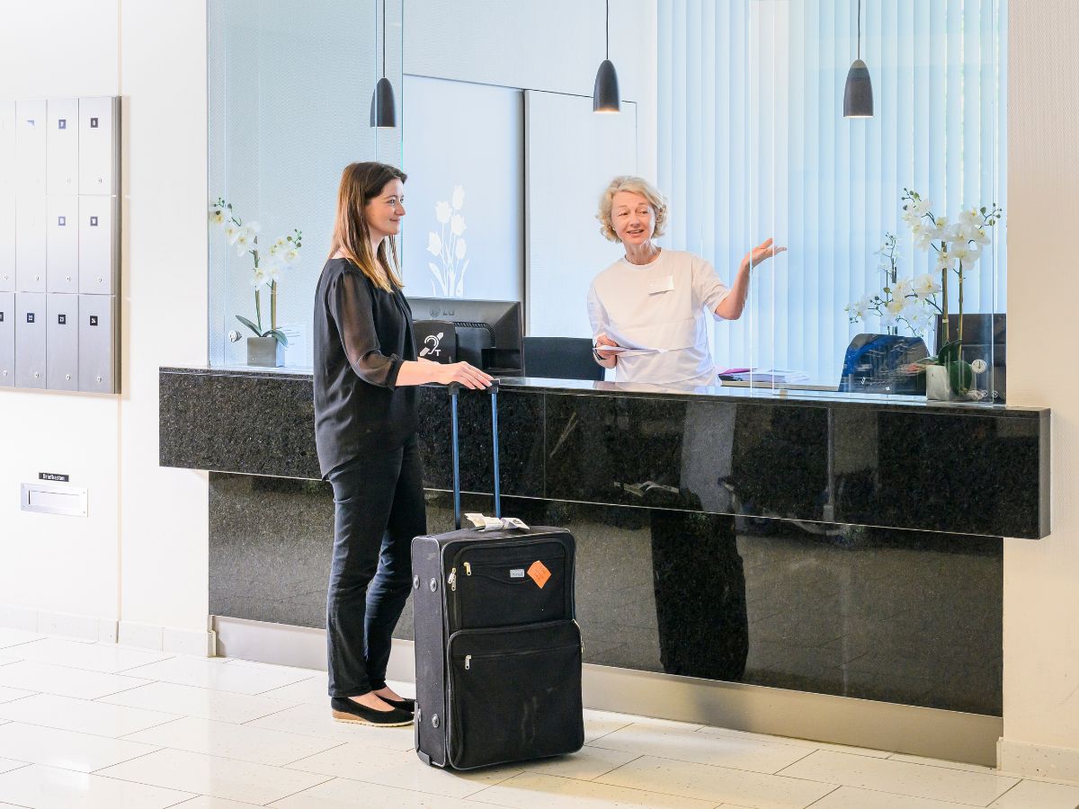 An der Rezeption zeigt die Rezeptionistin der Patientin mit Koffer den Weg