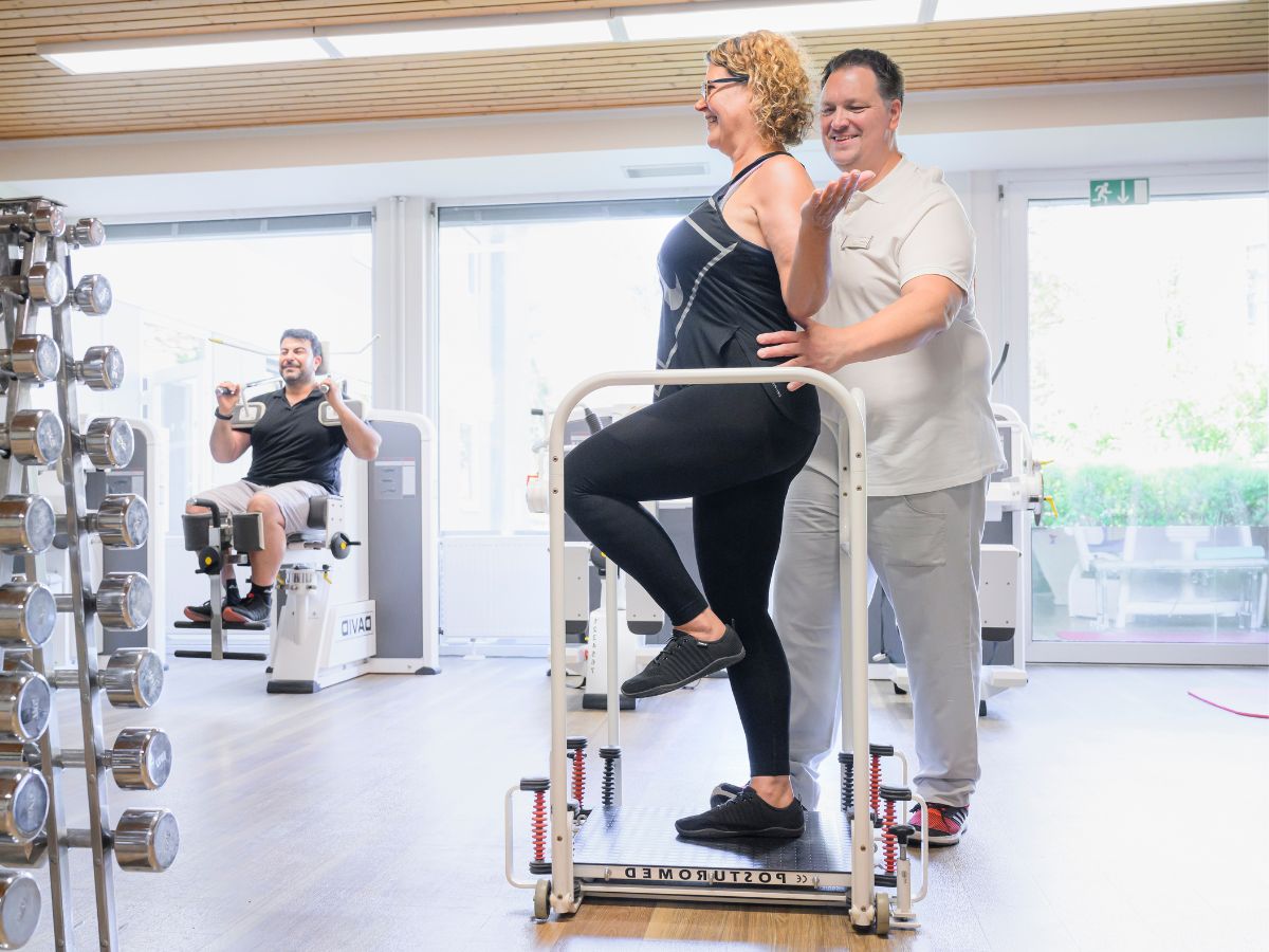 Therapeutin unterstützt Patientin auf Balance Board im Fitnessraum