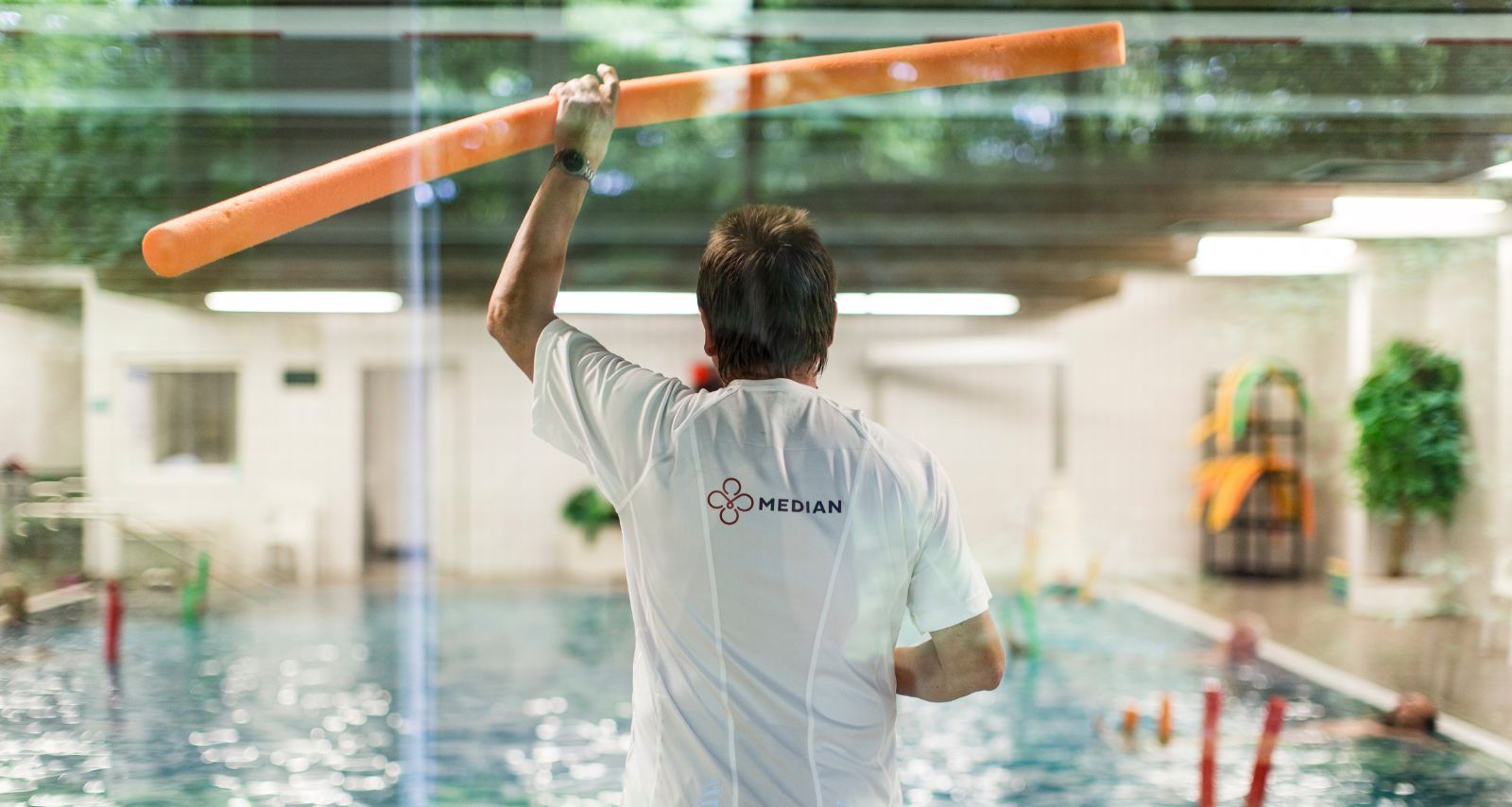 Therapeut hält am Beckenrand eine Poolnudel in der Hand und zeigt Patienten im Wasser die Übung.