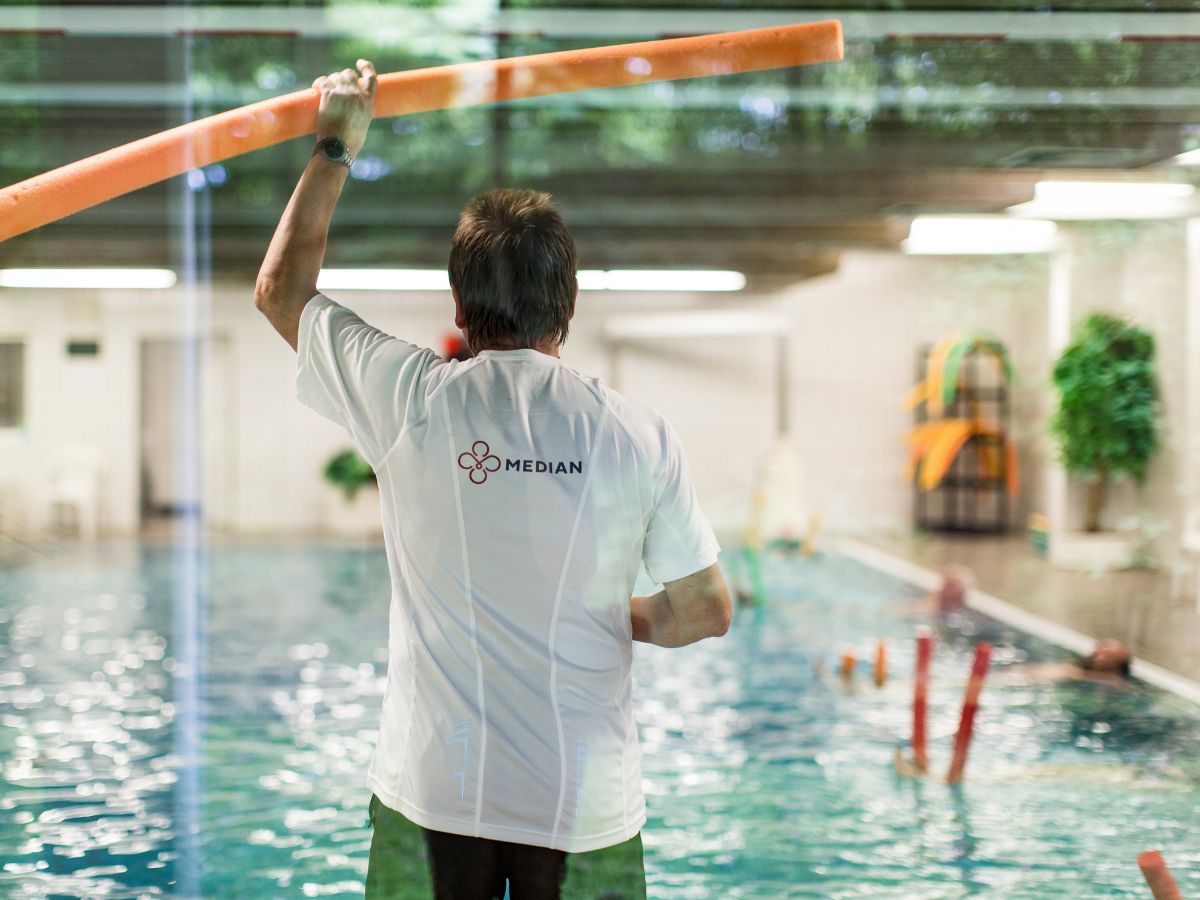 Therapeut hält am Beckenrand eine Poolnudel in der Hand und zeigt Patienten im Wasser die Übung.