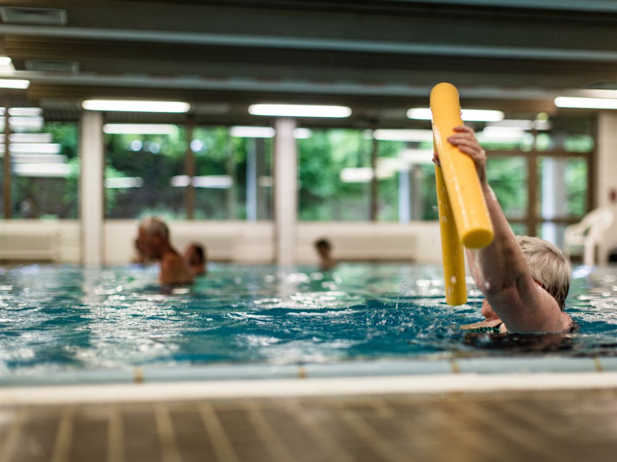 Im Wasserbecken hält eine Patientin eine Poolnudel hoch und macht Übungen 