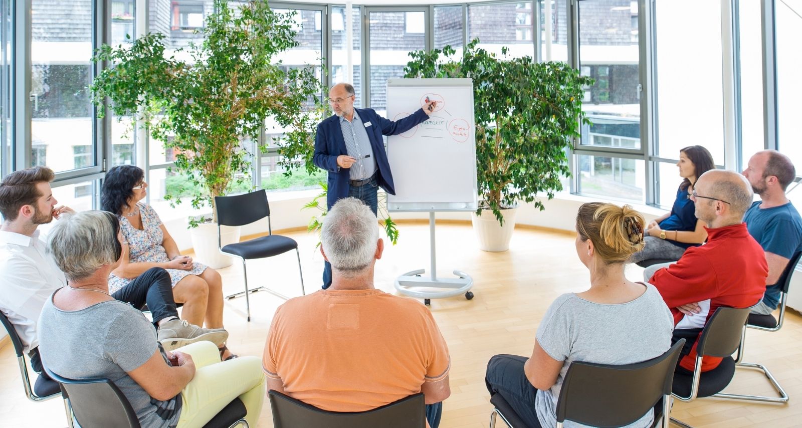 Personen die im Kreis sitzen und den Mann an der Tafel anschauen