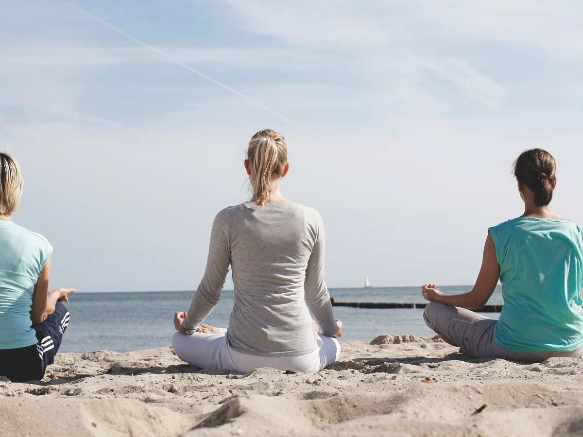 Drei Frauen sitzen im Schneidersitz am Strand und meditieren gemeinsam.