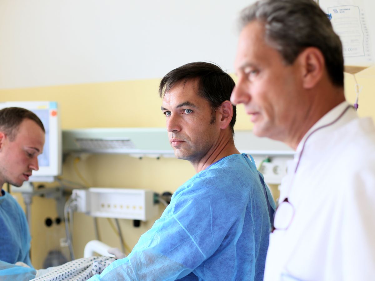 Ein Arzt und zwei Pfleger mit blauer Arbeitskleidung stehen am Patientenbett 