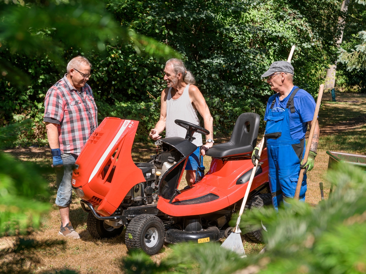 Drei Männer stehen im Garten an einem Aufsitzmäher mit geöffneter Motorhaube; einer hält einen Laubbesen.