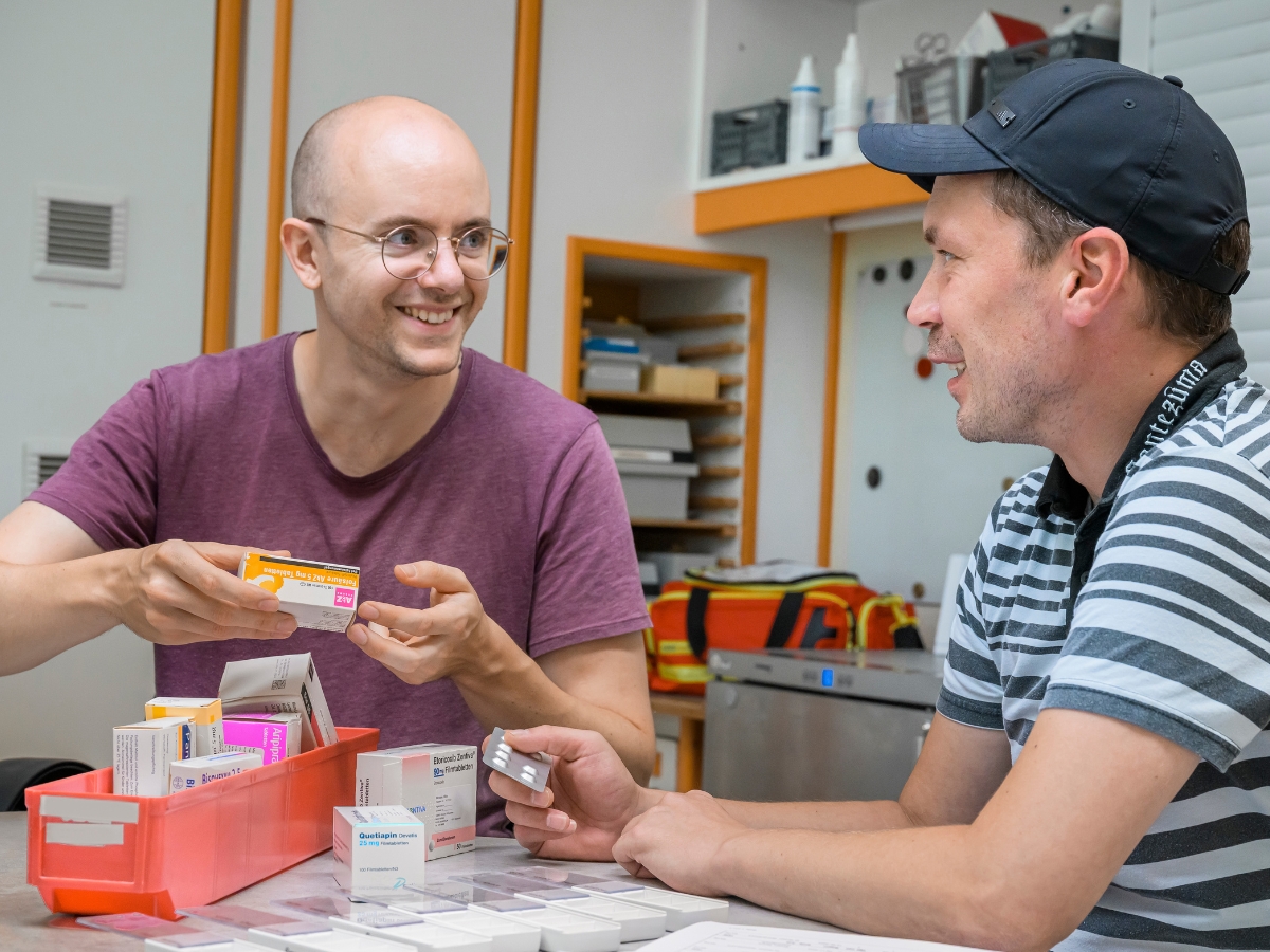 Zwei Männer besprechen Medikamente an einem Tisch mit Medikamentenschachteln und Dosierboxen.