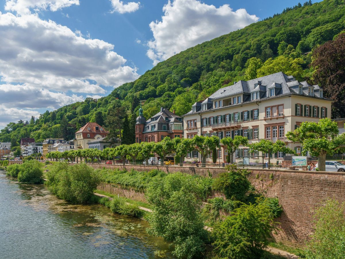 Fluss mit begrüntem Ufer und Häusern am Fuß eines bewaldeten Hangs