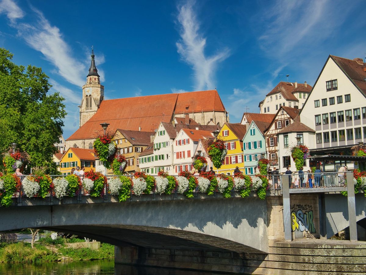 Bunte Häuser am Neckar in Tübingen, dahinter die Stiftskirche; eine blumengeschmückte Brücke im Vordergrund